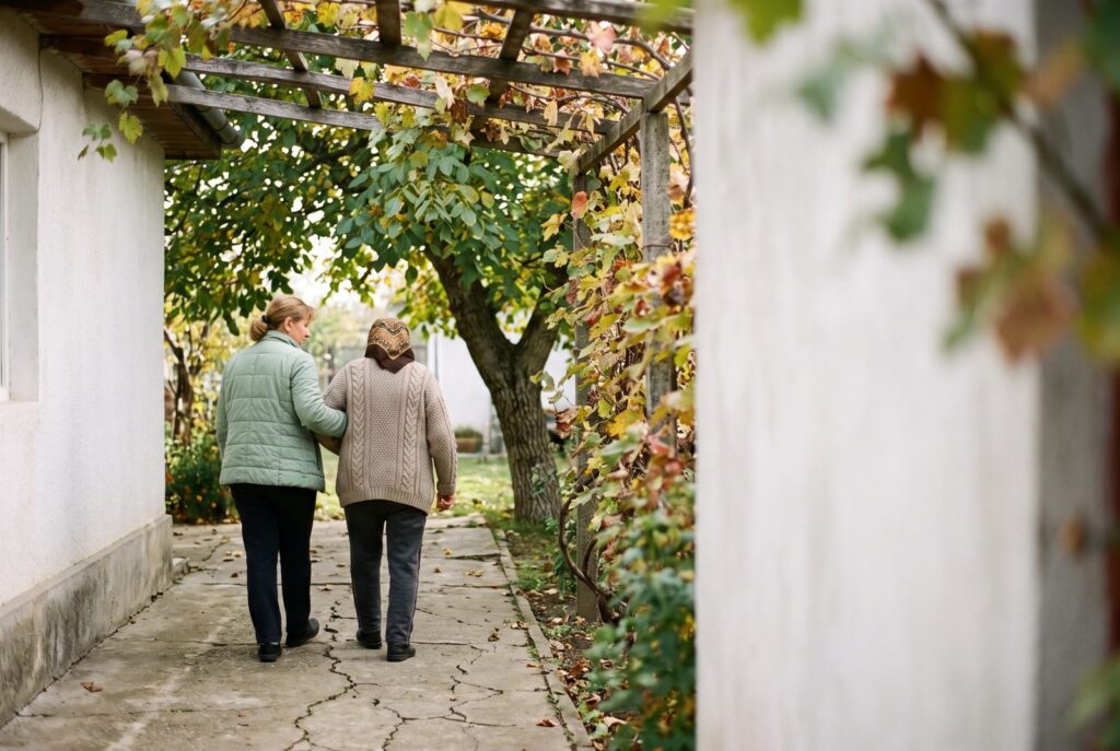 O femeie își ține mama de braț, pe o alee de grădină, sub o pergolă de viță, văzute din spate
