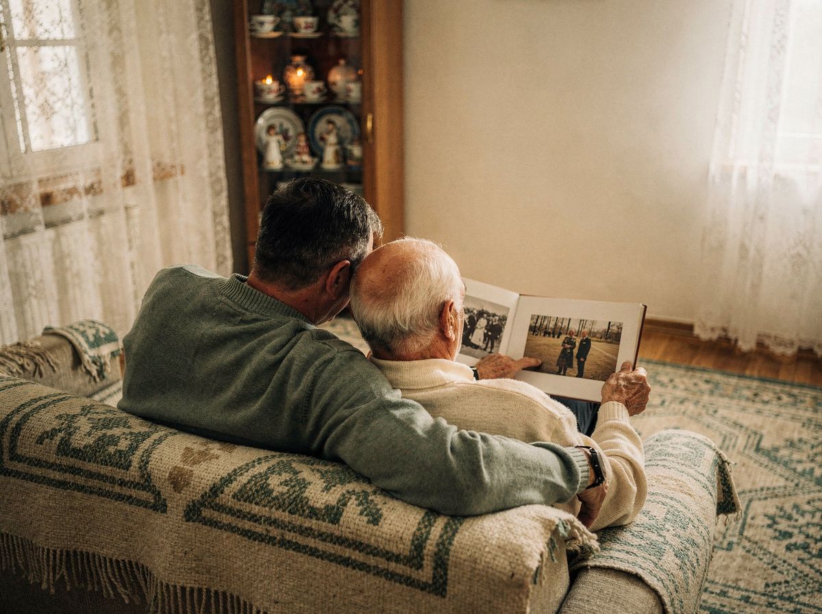 Un adult și un părinte vârstnic, văzuți din spate pe o canapea, privind împreună un album de fotografii