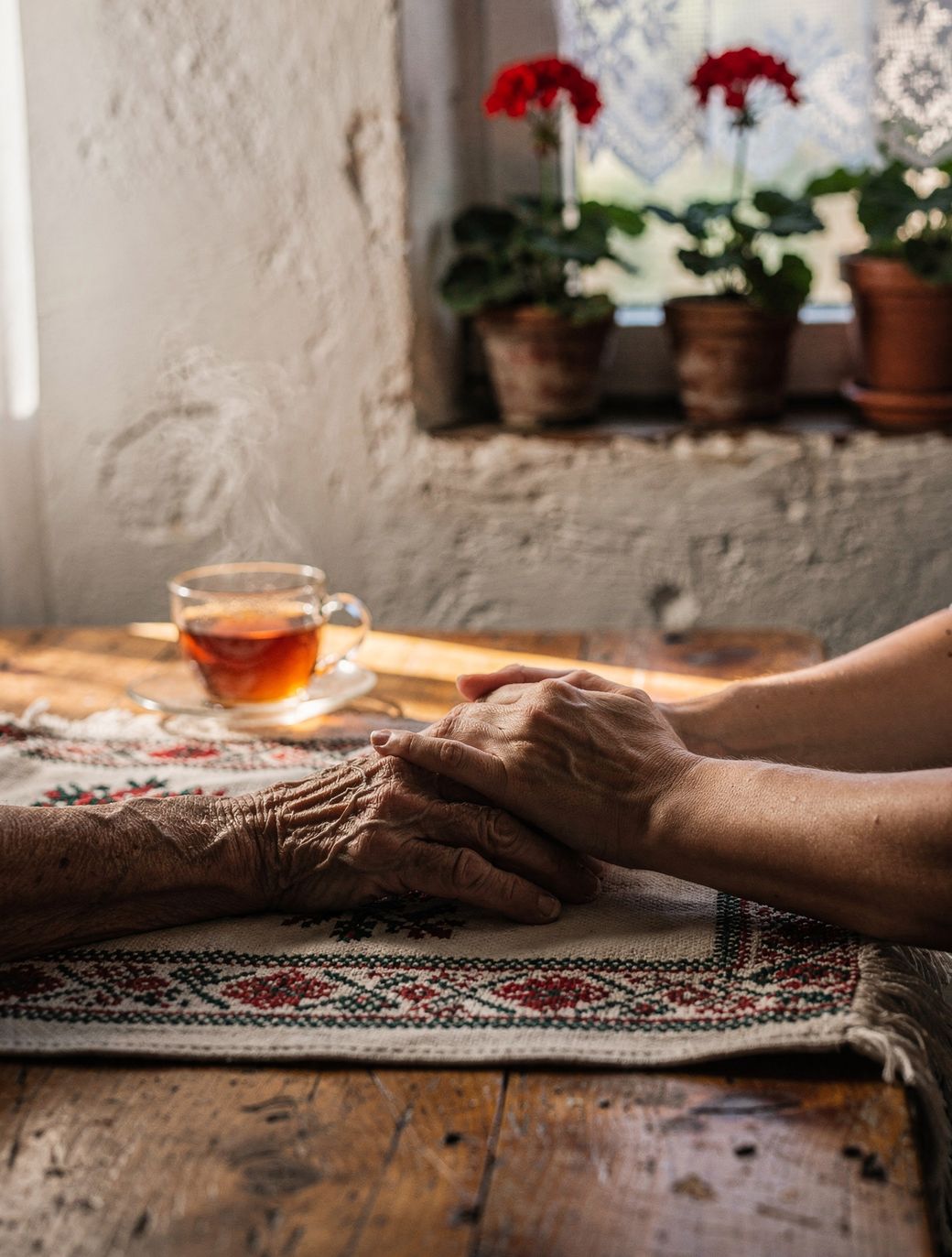 Mâinile împreunate ale unui adult și ale unui părinte vârstnic, pe o masă de bucătărie, lângă un ceai și mușcate la fereastră